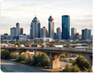 News thumbnail photograph of a modern city skyline viewed over a river valley or broad green foreground.